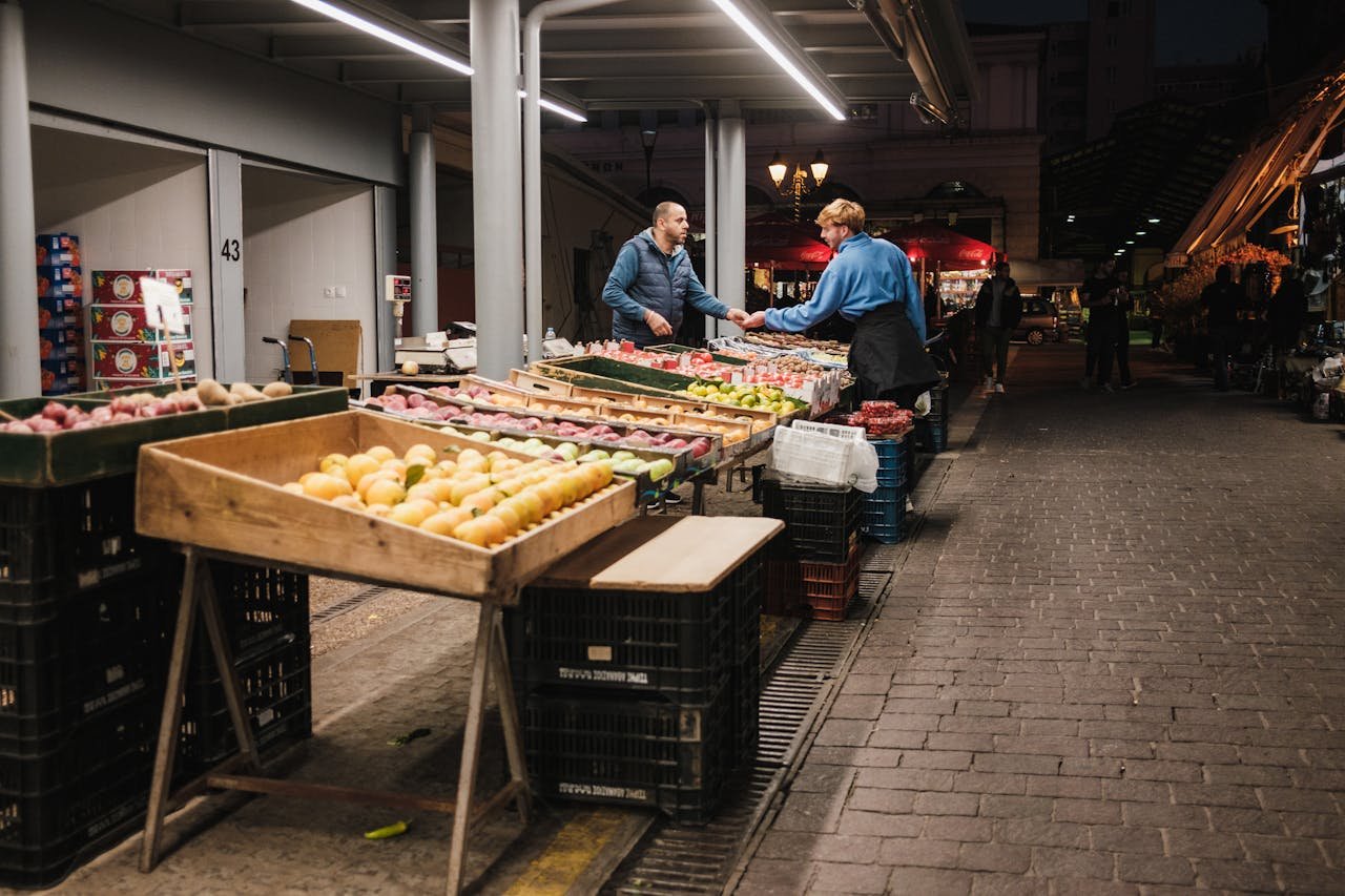 contact-img Two people exchanging goods at a well-lit night market in the city, surrounded by fresh produce.
