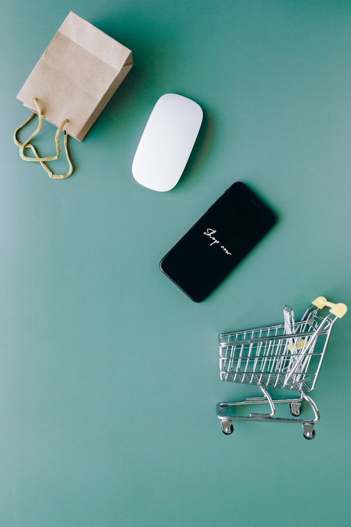 our-experience Top view of a smartphone, mouse, paper bag, and mini shopping cart on a green background, symbolizing e-commerce.